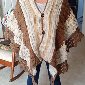 May include: A handmade crochet shawl in shades of brown, cream, and beige. The shawl features a V-neck design with two large brown buttons. The crochet pattern includes textured and openwork sections, creating a cozy and stylish garment.