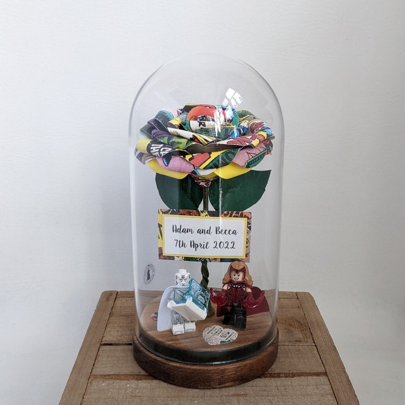 May include: A glass dome with a comic book paper rose inside. The rose is yellow, green, red, and blue. There are two small figurines at the base of the rose, one red and one blue. A sign at the base of the dome reads "Adam and Becca 7th April 2022".