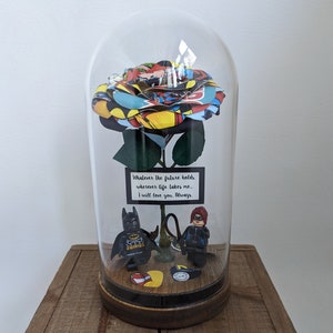 May include: A glass dome with a comic book themed rose inside. The rose is made of paper and has a green stem. There are two Lego minifigures at the base of the rose, one is Batman and the other is Batgirl. The text on the sign reads "Whatever the future holds, wherever life takes me... I will love you. Always."