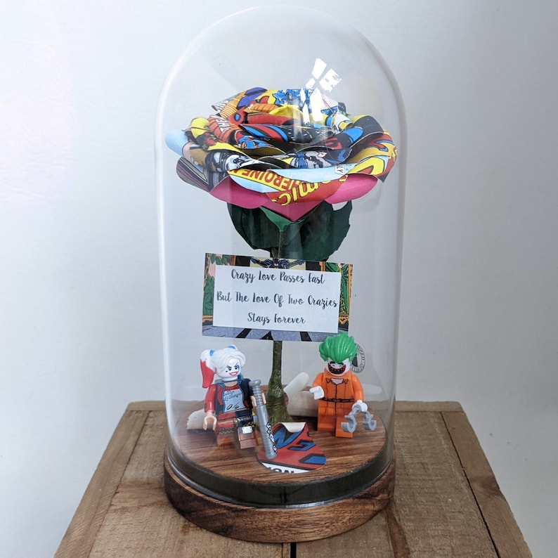 May include: A glass dome with a pink and yellow rose made of comic book pages. The rose is on a green stem with a sign that reads "Crazy love passes fast, but the love of two crazies stays forever". Two Lego figures, Harley Quinn and the Joker, are at the base of the rose.