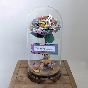 May include: A glass dome with a comic book rose inside. The rose is purple and has a green stem. A small Thanos Lego figure is at the base of the rose. The text on the card reads "You are my Endgame".