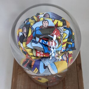 May include: A glass dome with a colorful paper rose inside. The rose is made from comic book pages with a superhero theme. The pages are in shades of red, yellow, blue, and green.