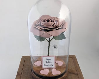 May include: A dusty rose leather rose flower inside a clear glass dome on a circular wooden base. The base features four small pink heart cutouts and a square white sign with black text that reads Happy Leather Anniversary.