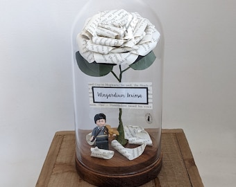 May include: A glass dome with a white paper flower made from book pages. The flower is on a green stem with a small figurine of a wizard holding a wand. The text on the label reads "Wingardium Leviosa".