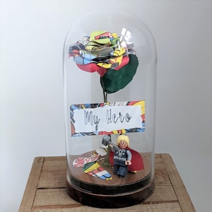 May include: A glass dome with a red, green, and yellow comic book rose inside. A Lego figure of Thor is standing on a wooden base with the words "My Hero" on a sign.