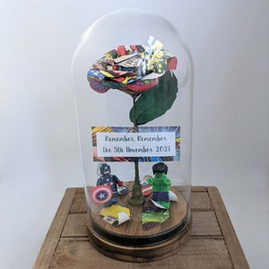 May include: A glass dome with a red, green, and yellow paper rose inside. The rose is made from comic book pages. There are two small superhero figurines at the base of the rose, one is Captain America and the other is the Hulk. A small card at the base of the rose reads "Remember, Remember the 5th November 2021."