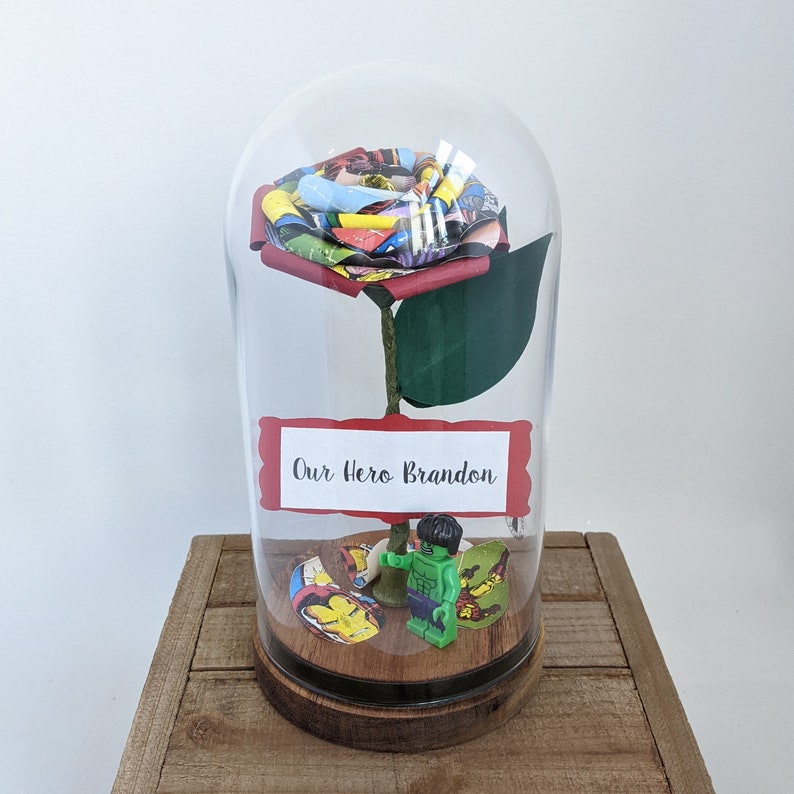 May include: A glass dome with a red rose made of comic book pages. The rose has a green stem and leaves. A small green figurine of the Hulk is at the base of the rose. A white label on the dome reads "Our Hero Brandon".