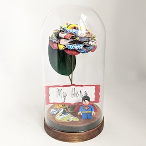 May include: A glass dome with a red and yellow comic book rose inside. The rose is on a green stem and there is a small Superman Lego figure at the base of the rose. A red sign with the words "My Hero" is in front of the rose.