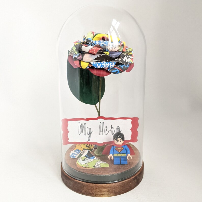 May include: A glass dome with a red and yellow comic book rose inside. The rose is on a green stem and there is a small Superman Lego figure at the base of the rose. A red sign with the words "My Hero" is in front of the rose.