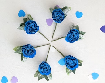 May include: Six handmade blue paper roses with green leaves and stems arranged in a circular pattern. The roses are surrounded by blue and purple heart-shaped confetti on a white surface.
