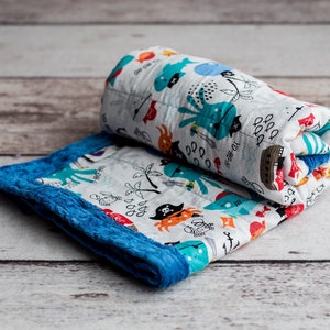 May include: A white and blue baby blanket with a pirate theme. The blanket features a variety of colorful illustrations, including an octopus, a shark, a pirate, and a treasure chest. The blanket is rolled up and has a blue velvet border.