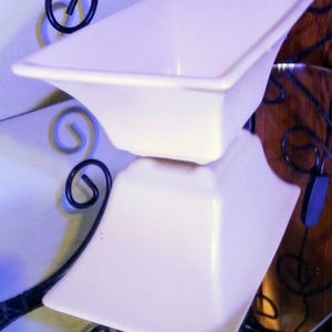 May include: A white ceramic bowl with a square, flared rim and a tapered base. The bowl is positioned on a reflective surface, creating a clear mirror image of the object below it. The design features smooth, clean lines and a glossy finish.