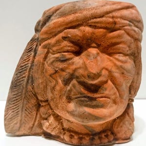 May include: A terracotta-colored ceramic mug shaped like the head of a person wearing a feathered headdress. The sculpture features detailed facial expressions, including closed eyes, a prominent nose, and a textured headband with a feather.