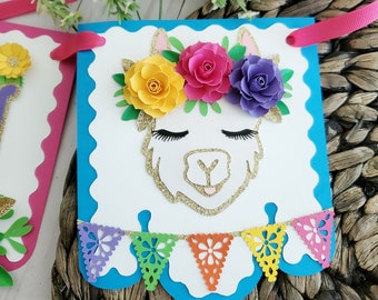 May include: A blue and white llama party banner with a llama face and a flower crown. The banner has a scalloped edge and a string of colorful paper flags.
