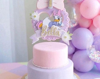 May include: A two-tiered cake with pink and lavender frosting, decorated with a Daisy Duck cake topper. The topper features a pink bow, the number 1, and the name "Bella" in gold glitter. The cake sits on a wooden base.
