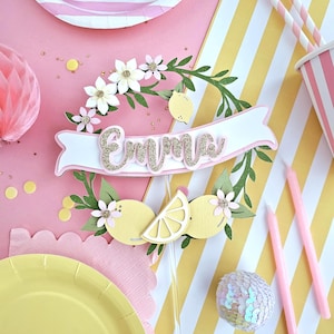 May include: A party scene in pink and yellow. A cake topper with the name "Emma" in gold glitter is surrounded by lemon and floral decorations. Pink plates, cups, straws, and candles complement the theme, along with decorative balls. A cheerful and bright setting.