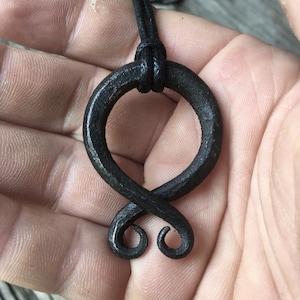 May include: A black metal pendant in the shape of a stylized knot, hanging from a black leather cord.