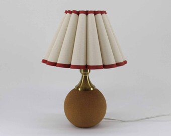 May include: A small table lamp with a round, brown base and a brass-colored stem. The lampshade is cream-colored with a pleated design and a red trim. The lamp is plugged into a white cord.