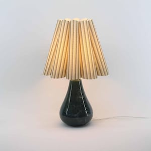 Puede incluir: Lámpara de mesa decorativa con base en forma de lágrima verde oscuro y pantalla a rayas. La pantalla tiene rayas verticales en beige y gris oscuro. La lámpara está encendida, emitiendo un brillo cálido.