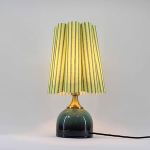 Könnte beinhalten: Eine Tischlampe mit einem grün-schwarzen Sockel, einem messingfarbenen Hals und einem gelben Lampenschirm mit vertikalen schwarzen Streifen. Die Lampe ist mit einem schwarzen Kabel angeschlossen. Der Lampenschirm hat ein plissiertes Design.