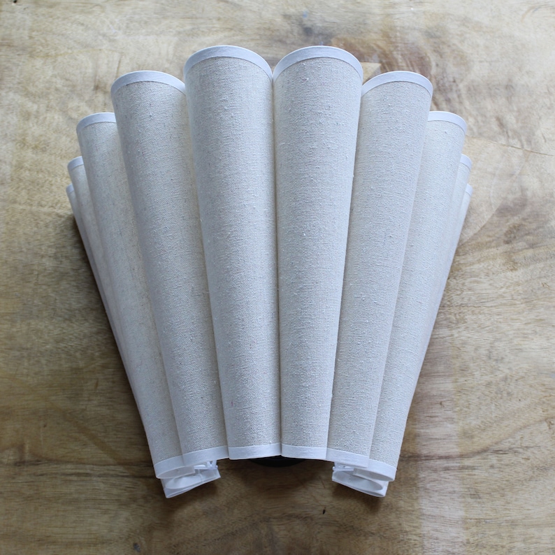 May include: A cream-colored lampshade with a pleated design. The shade is made of a textured fabric and features a white trim along the top and bottom edges. The shade is cone-shaped and has a fan-like appearance.