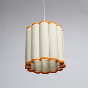 May include: A pendant light fixture with a cylindrical lampshade. The shade is made of a cream-colored fabric with vertical lines and is trimmed with orange bands at the top and bottom. The light hangs from a white cord.