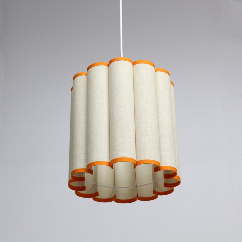 May include: A pendant light fixture with a cylindrical lampshade. The shade is made of a cream-colored fabric with vertical lines and is trimmed with orange bands at the top and bottom. The light hangs from a white cord.