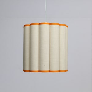 May include: A cream-colored pendant light with a cylindrical, scalloped shade. The shade has a textured fabric appearance and is trimmed with bright orange bands at the top and bottom. The light hangs from a white cord.