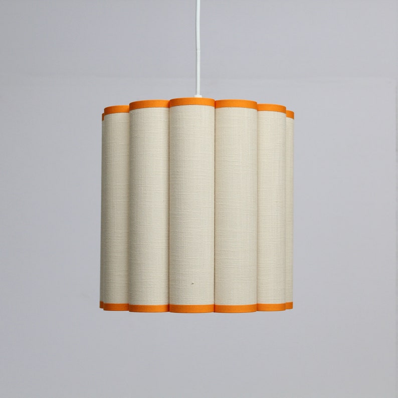 May include: A cream-colored pendant light with a cylindrical, scalloped shade. The shade has a textured fabric appearance and is trimmed with bright orange bands at the top and bottom. The light hangs from a white cord.