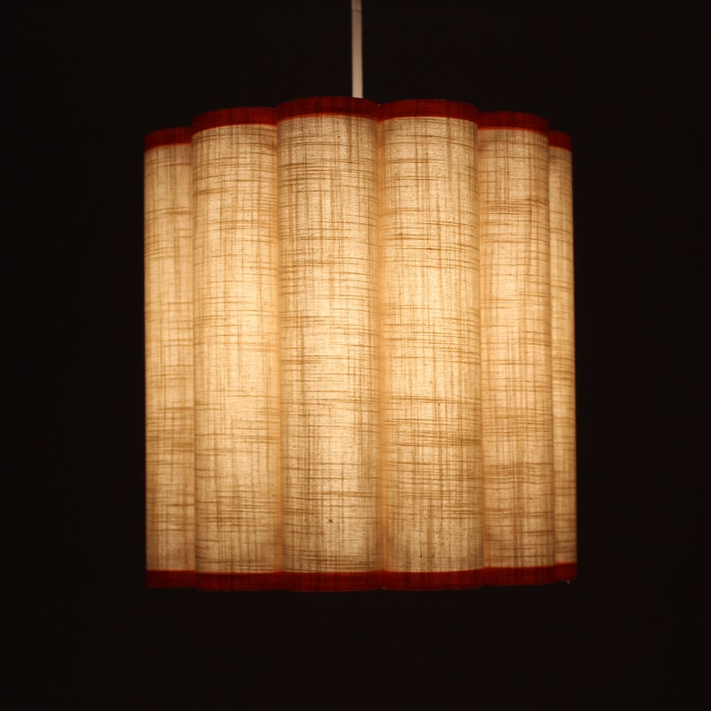 May include: A cylindrical pendant light fixture with a textured, beige fabric shade. The shade has a scalloped top and bottom, trimmed with a thin, red border. The light fixture hangs from a white cord against a dark background.