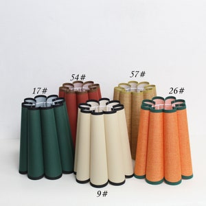 May include: Five different lampshades with pleated fabric in various colors, including green, beige, orange, brown, and tan. The lampshades are arranged in a row on a white surface. The numbers 17#, 54#, 57#, 26#, and 9# are written above the lampshades.