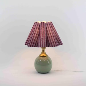 Può includere: Lampada da tavolo decorativa con paralume plissettato a righe verticali viola, rosa e nere. Ha una base in ottone e un corpo rotondo in ceramica verde salvia. L'altezza è di circa 30 cm.