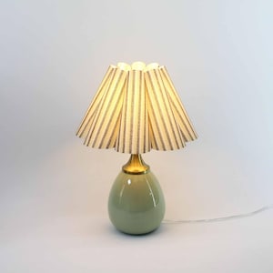 Könnte beinhalten: Eine kleine, dekorative Tischlampe mit einem hellgrünen, eiförmigen Sockel und einem plissierten Lampenschirm. Der Lampenschirm ist cremefarben mit vertikalen braunen Streifen. Die Lampe hat einen messingfarbenen Hals und ein weißes Netzkabel.