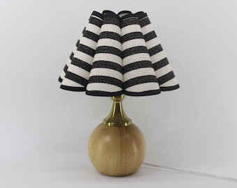 Op de afbeelding: Een kleine tafellamp met een ronde houten voet en een messingkleurige steel. De lampenkap is zwart-wit gestreept, met een geplooid ontwerp. De lamp staat op een witte achtergrond.
