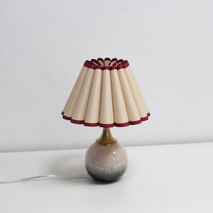 May include: A table lamp with a pleated beige lampshade with a burgundy trim. The lamp base is a speckled ceramic sphere in shades of gray and pink.