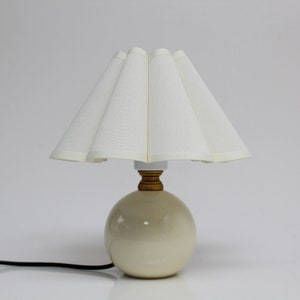 May include: A white ceramic table lamp with a pleated white fabric lampshade. The lamp has a round base and a gold-colored metal accent.