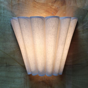 May include: A wall-mounted light fixture with a pleated, off-white fabric shade. The shade has a fan-like design with a light blue trim. The fixture is mounted on a wooden surface, creating a warm, inviting ambiance.