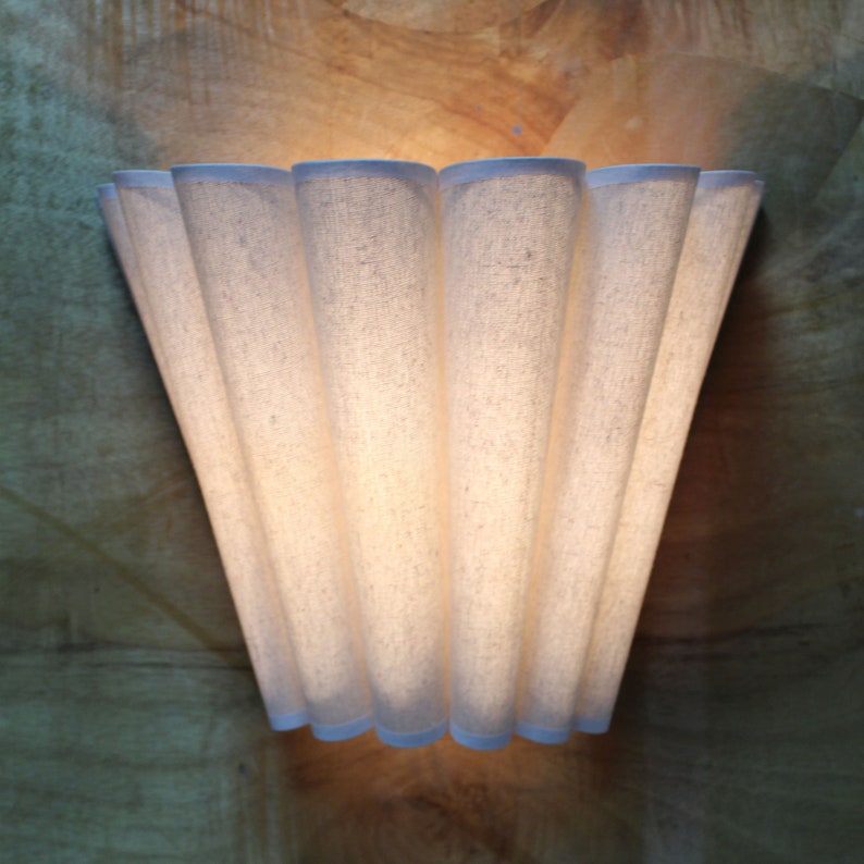 May include: A wall-mounted light fixture with a pleated, off-white fabric shade. The shade has a fan-like design with a light blue trim. The fixture is mounted on a wooden surface, creating a warm, inviting ambiance.