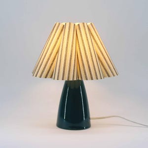 May include: A dark teal ceramic table lamp with a striped lampshade. The lampshade has vertical stripes in beige and dark brown. The lamp has a rounded base and a conical shape. The lamp is plugged into a white wall.