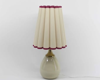 Puede incluir: Una lámpara de mesa color crema con una pantalla plisada. La pantalla tiene un ribete burdeos en la parte superior e inferior. La base es de color beige claro con un cuello color latón.