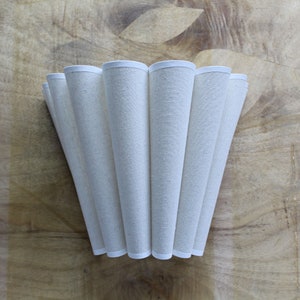May include: Cream-colored lampshade with a pleated design. The shade is made of a textured fabric and has a white trim along the top and bottom edges. The lampshade is set against a wooden background.
