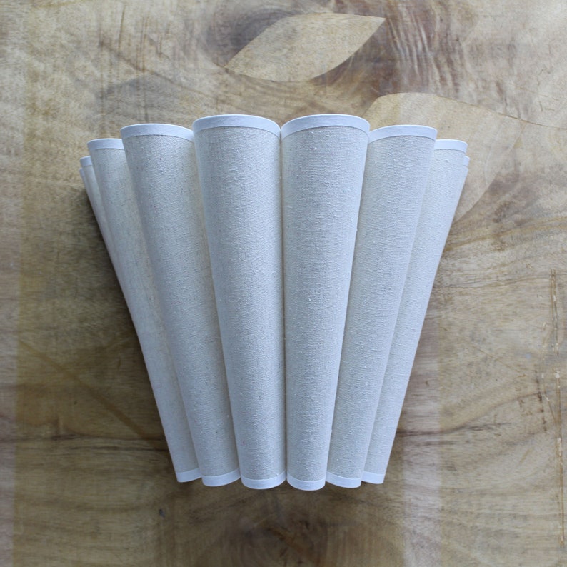May include: Cream-colored lampshade with a pleated design. The shade is made of a textured fabric and has a white trim along the top and bottom edges. The lampshade is set against a wooden background.