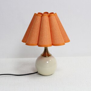 May include: A vintage table lamp with a round, cream-colored base and a brass neck. The lampshade is orange with a textured fabric and a scalloped design. A black power cord extends from the base.