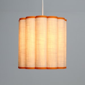 May include: A cylindrical pendant light fixture with a white linen shade and orange trim. The shade is composed of vertical, fluted panels. The light hangs from a white cord, casting a warm glow.
