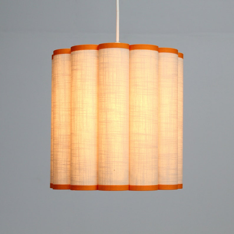 May include: A cylindrical pendant light fixture with a white linen shade and orange trim. The shade is composed of vertical, fluted panels. The light hangs from a white cord, casting a warm glow.