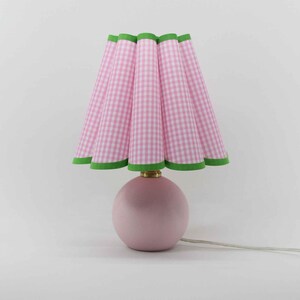 May include: A pink table lamp with a round base and a pleated lampshade. The lampshade features a pink and white gingham pattern with green trim. The lamp has a brass-colored neck and a white cord.