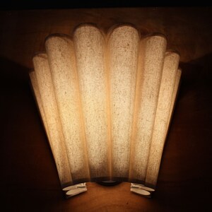 May include: A wall-mounted light fixture with a fan-shaped design. The fixture has seven vertical, cylindrical shades made of a textured, off-white fabric. The light shines through the shades, creating a warm glow against the dark brown wall.