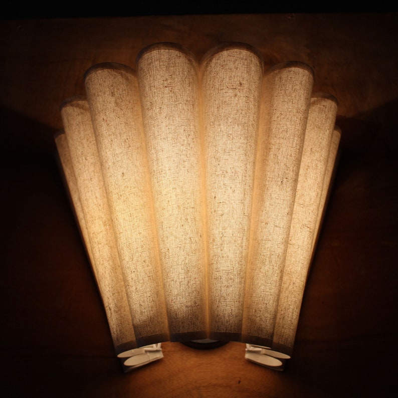 May include: A wall-mounted light fixture with a fan-shaped design. The fixture has seven vertical, cylindrical shades made of a textured, off-white fabric. The light shines through the shades, creating a warm glow against the dark brown wall.