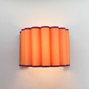 May include: An orange wall sconce with a cylindrical design. The sconce has a series of vertical, tube-like shades with a dark purple trim. The light fixture is mounted on a white wall.