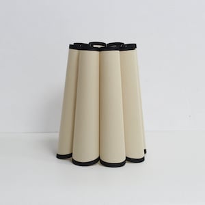 May include: A cream-colored lampshade with a black trim. The lampshade is pleated and has a rounded shape.
